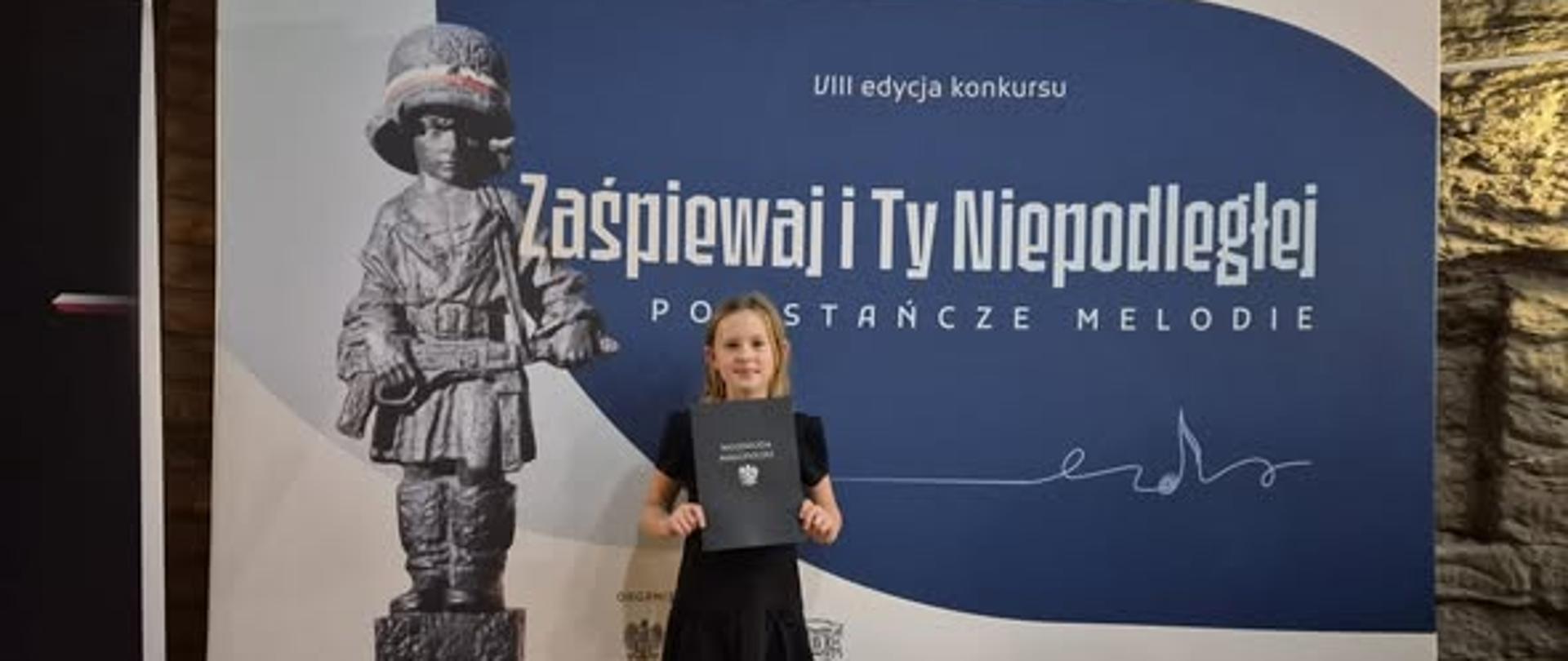 Karolina pozuje do zdjęcia z dyplomem, w tle baner przedstawiający rzeźbę małego chłopca w hełmie z karabinem, na niebieskim tle widnieje napis: "VII edycja konkursu Zaśpiewaj i Ty Niepodległej Powstańcze melodie"