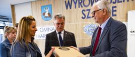 Minister Piechowiak wręczający komputer kobiecie, w tle burmistrz Janusz Piechocki i 2 inne osoby, za nimi ściana z herbem gminy Margonin, napisem Gmina Margonin Wyrównujemy szanse