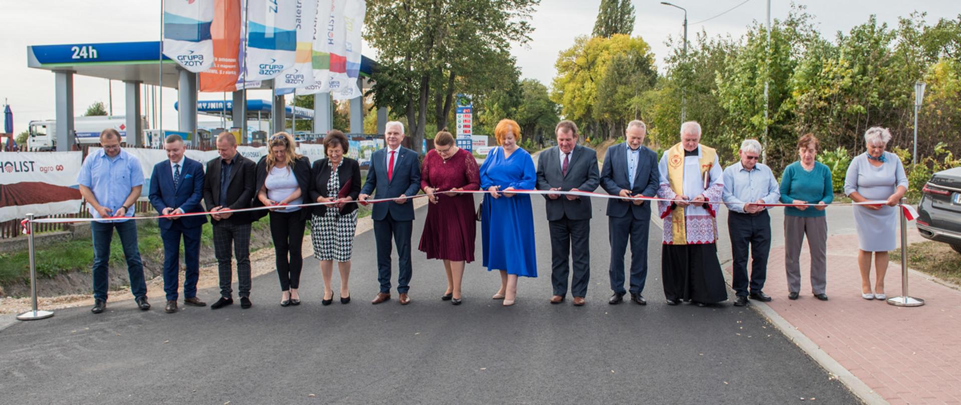 Moment przecięcia wstęgi. Na fotografii widzimy stojących w szeregu w poprzek drogi uczestników w trakcie rozpostartej przez drogę ma dwóch metalowych słupkach biało - czerwonej wstęgi.