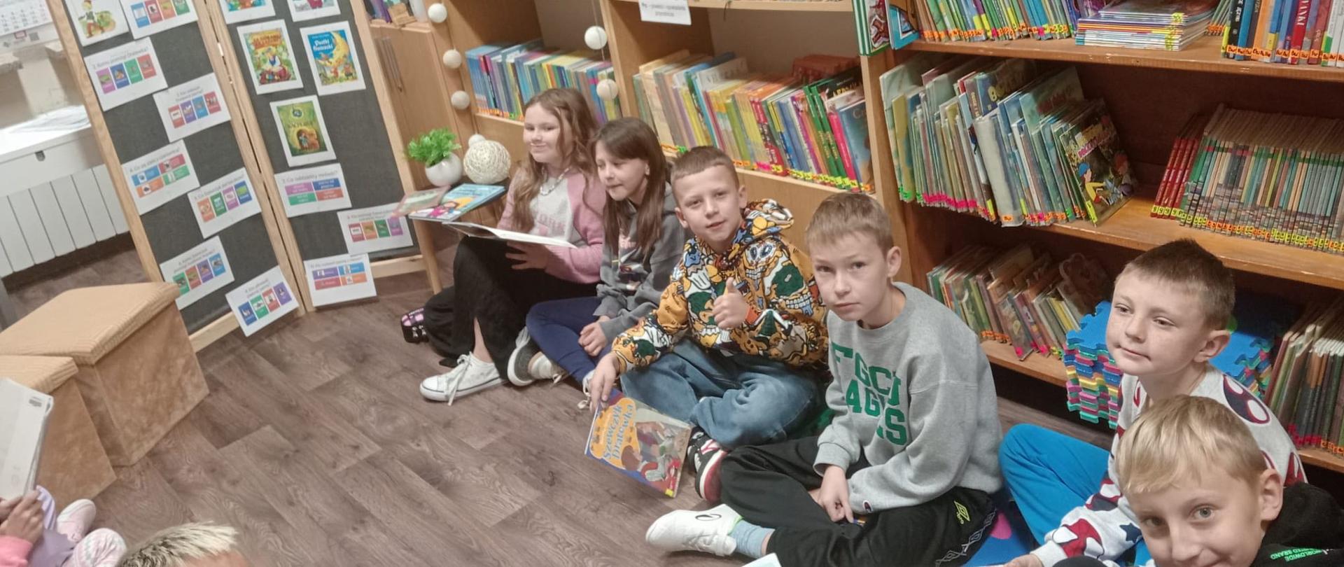Dzieci z klasy 3 siedzą na piankowych matach w bibliotece w kręgu naprzeciw siebie, każdy z nich czyta głośno po fragmencie książki, po lewej i prawej stronie za dziećmi półki z książkami
