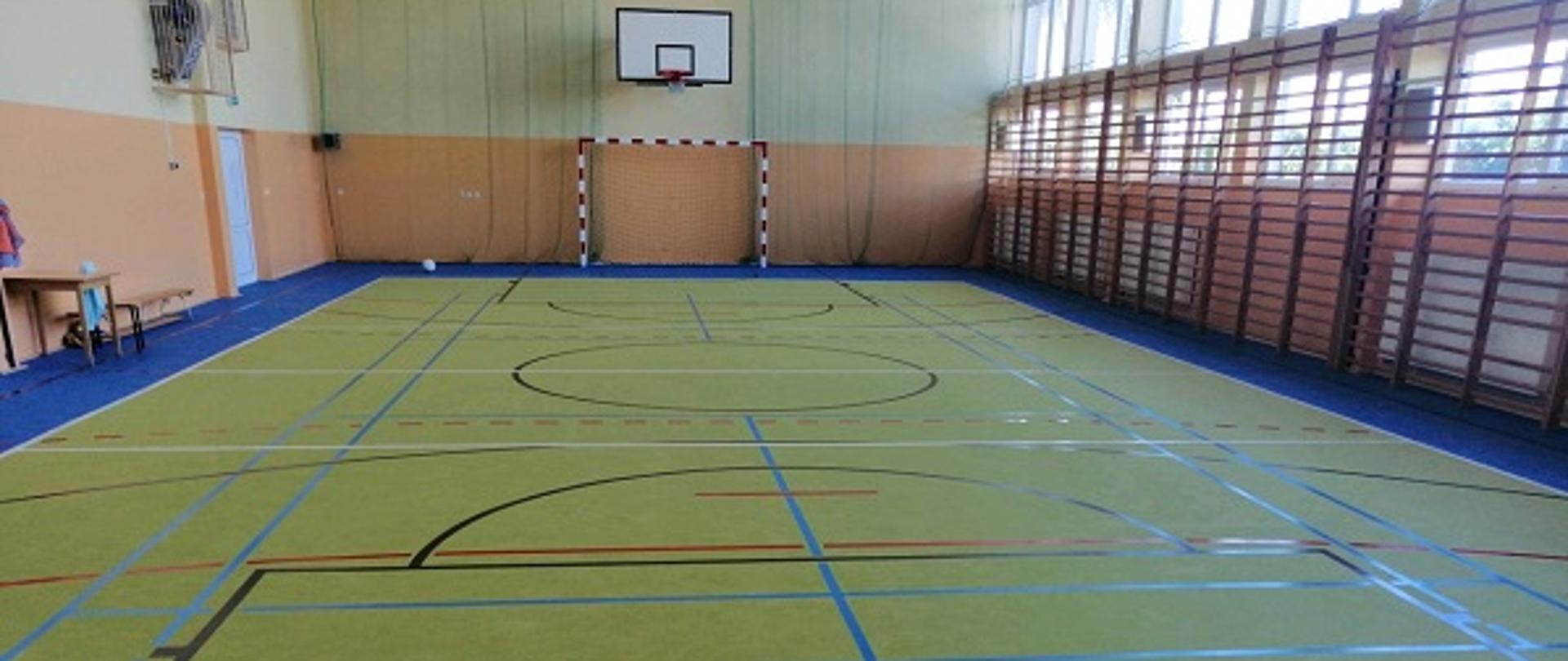Na zdjęciu widoczna szkolna sala gimnastyczna z wyremontowaną podłogą sportową w kolorze żółtym i niebieskim. Na wprost widoczna bramka i kosz. Po prawej stronie znajdują się okna i drabinki gimnastyczne.