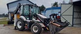 Koparko-ładowarka Hidromek 102B na placu, z przymocowaną przednią łyżką chwytającą drewniane elementy. W tle widoczne są budynki przemysłowe i zbiorniki.