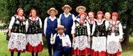 do zdjecia na zielonej trawie pozują kobiety w ludowych strojach czerwonych spódnicach białych haftowanych fartuszkach i czarnych gorsetach oraz mężczyźni w granatowych spodniach i kamizelkach i białych koszulach 