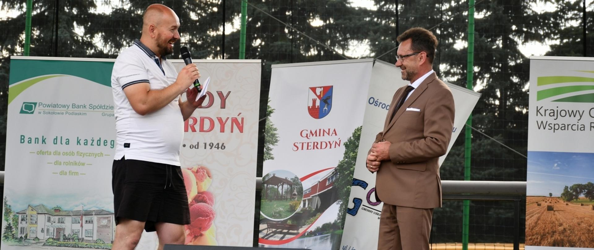 Na zdjęciu na scenie z lewej strony prezes stowarzyszenia "Piłkarskie siódemki" dziękuje staroście sokołowskiemu za wsparcie turnieju. W tle stoją rollupy instytucji.
