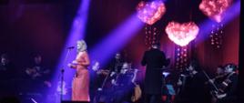 Wokalistka Katarzyna Żak śpiewa na scenie, za nią Sądecka Orkiestra Kameralna w składzie 10 osób z dyrygentem Leszkiem Mieczkowskim.