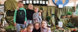 W sali lekcyjnej pod oknem i ścianą stoją stoły nakryte zielonym płótnem, na nich umieszczone zostały rośliny w doniczkach z opisami oraz doświadczenia związane z wodą, po lewej stronie z niebieskiego płótna udrapowany wodospad, nad min wykonany z papieru i sznurka schemat obiegu wody składający się z chmurek deszczowych i kropel, na środku stoi trzech chłopców, przed nimi siedzą 3 dziewczynki