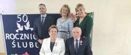 Państwo Ewa i Wiesław na tle granatowej tablicy z napisem wraz z Wójtem, Z-cą Wójta i Z-cą Kierownika USC