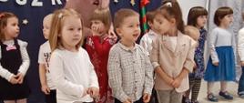 Przedszkolaki stoją w sali na tle ścianki "Pasowanie na przedszkolaka". 