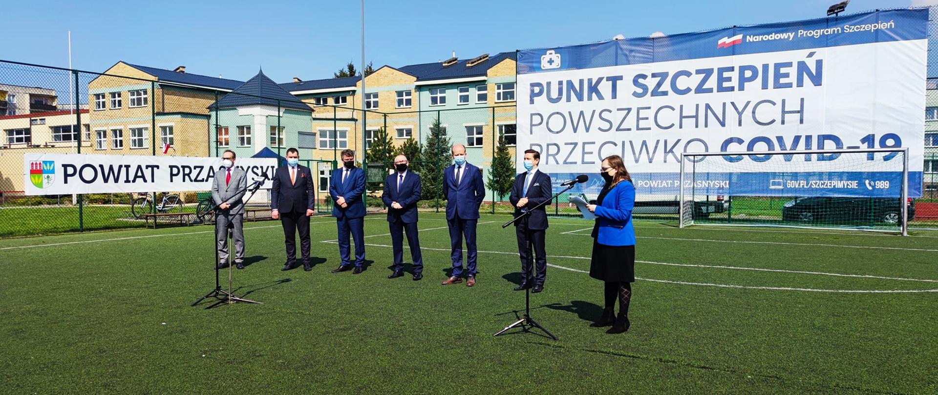 Zdjęcie przedstawia konferencję prasową z udziałem Wojewody Mazowieckiego w dniu otwarcia pilotażowego punktu szczepień w Przasnyszu. W pierwszym planie od lewej: Przewodniczący Rady Powiatu Przasnyskiego Paweł Szczepkowski, Dyrektor Samodzielnego Publicznego Zespołu Zakładów Opieki Zdrowotnej w Przasnyszu Grzegorz Magnuszewski, Sekretarz Stanu w Ministerstwie Spraw Wewnętrznych i Administracji Maciej Wąsik, Starosta Przasnyski Krzysztof Bieńkowski, Wojewoda Mazowiecki Konstanty Radziwiłł, Dyrektor Mazowieckiego Oddziału Wojewódzkiego NFZ Zbigniew Terek, prowadząca konferencję pracownica Delegatury Mazowieckiego Urzędu Wojewódzkiego. W tle siatka boiska na której umieszczono dwa banery: po lewej o treści Powiat Przasnyski, a po prawej o treści Narodowy Program Szczepień, Punkt Szczepień Powszechnych Przeciwko Covid-19. Za siatką prezentuje się Zespół Szkół Powiatowych przy ulicy Sadowej, gdzie na parterze zlokalizowano punkt szczepień.