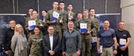 Zdjęcie grupowe. Zwycięzcy zawodów w mundurach stoją na podium z pucharami i dyplomami. Wraz z nimi opiekunowie i organizatorzy.