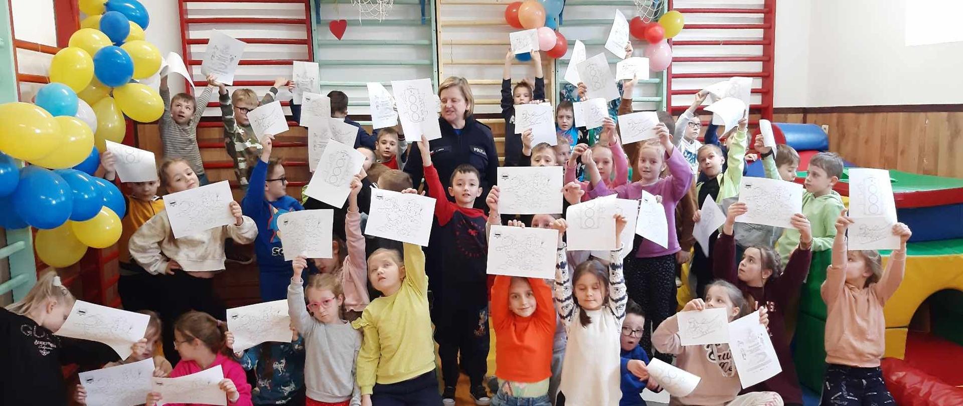 Pani dzielnicowa wraz z dziećmi na tle kolorowych balonów, ustawiona pod drabinkami. Dzieci w dłoniach trzymają malowanki " Bezpieczne ferie zimowe" 