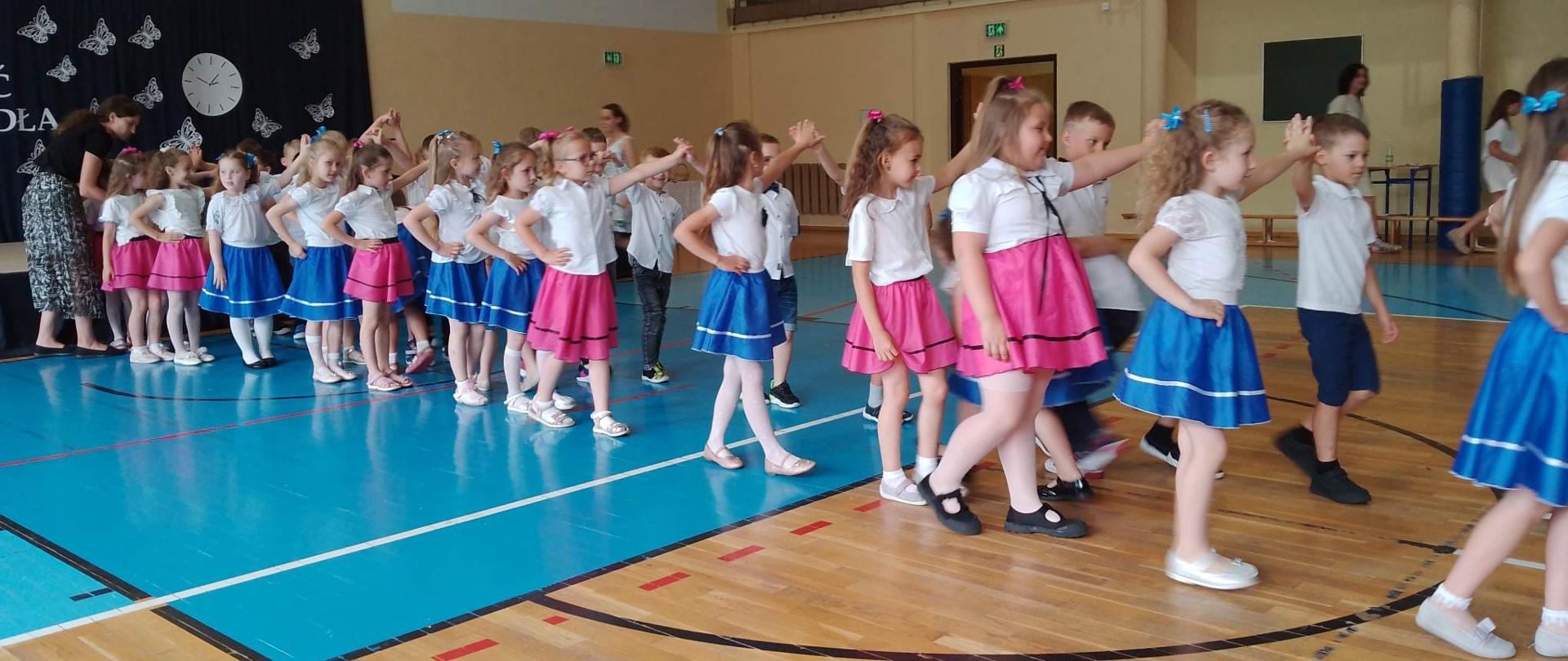 Dziewczynki w niebieskich si różowych spódniczkach oraz białych bluzkach tańczą w parach poloneza z chłopcami w białych koszulach i ciemnych spodniach. 