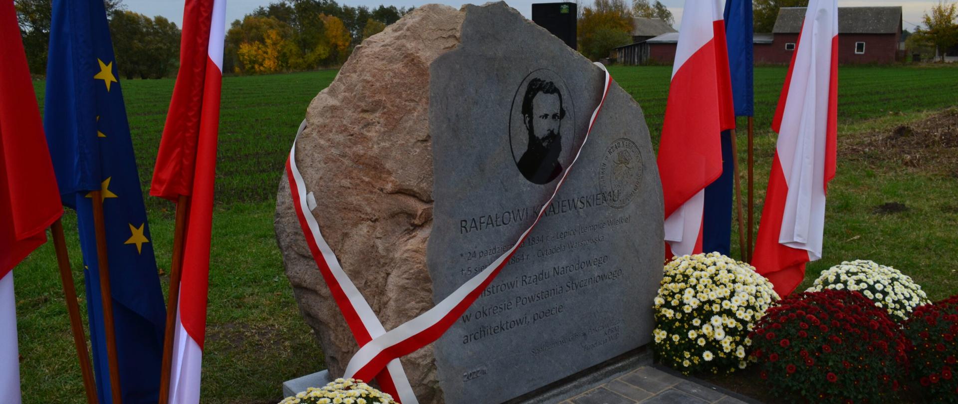 Pomnik Rafała Krajewskiego w Łępicach – kamień z portretem i inskrypcją, ozdobiony biało-czerwoną wstęgą, otoczony flagami Polski i UE oraz kwiatami. W tle widoczne pole, drzewa i budynki.
