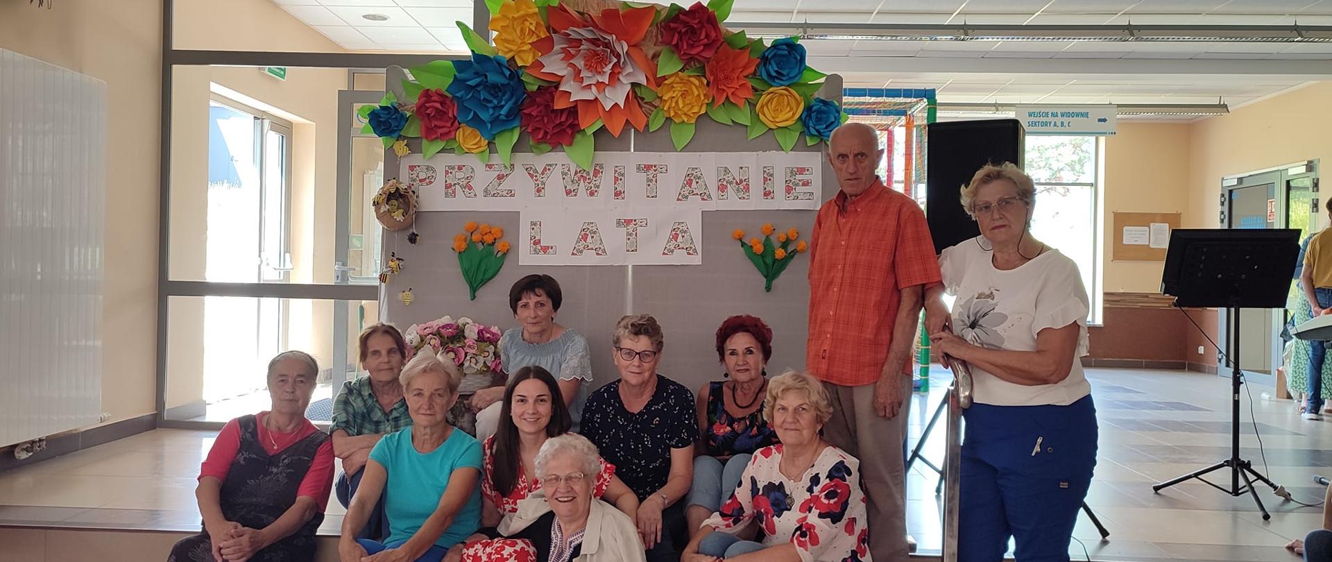 Członkowie Klubu Senior+ w hali widowiskowo-sportowej LEGA w Olecku. W tle tablica z napisem przywitanie lata. 