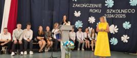 Kobieta stoi za mównicą i lewą ręką trzyma mikrofon. Obok niej stoi kobieta w zółtej sukience. Za nimi siedzą dzieci na ławkach. Napis na ścianie Zakończenie roku szkolnego 2024-2025