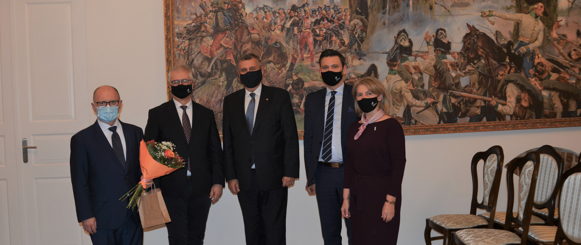 Jedna kobieta i czterech mężczyzn w maseczkach stoją na tle obrazu we wnętrzu odrestaurowanego budynku Austerii Raszyńskiej