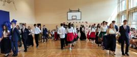 Uczniowie klasy ósmej tańczą poloneza: rozchodzą się parami na prawo i lewo, ubrani są na galowo, dziewczynki mają długie kwieciste spódnice