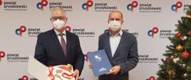 Krzysztof Rymuza Starosta Pruszkowski i Grzegorz Kamiński Członek Zarządu Powiatu Pruszkowskiego obok przystrojonej choinki. W ręku trzymają upominki dla ostatniego dziecka, które przyszło na świat w szpitalu powiatowym w 2020 roku.