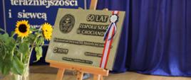 Odsłonięta tablica pamiątkowa, żółto-czarne kwiaty w wazonie. 