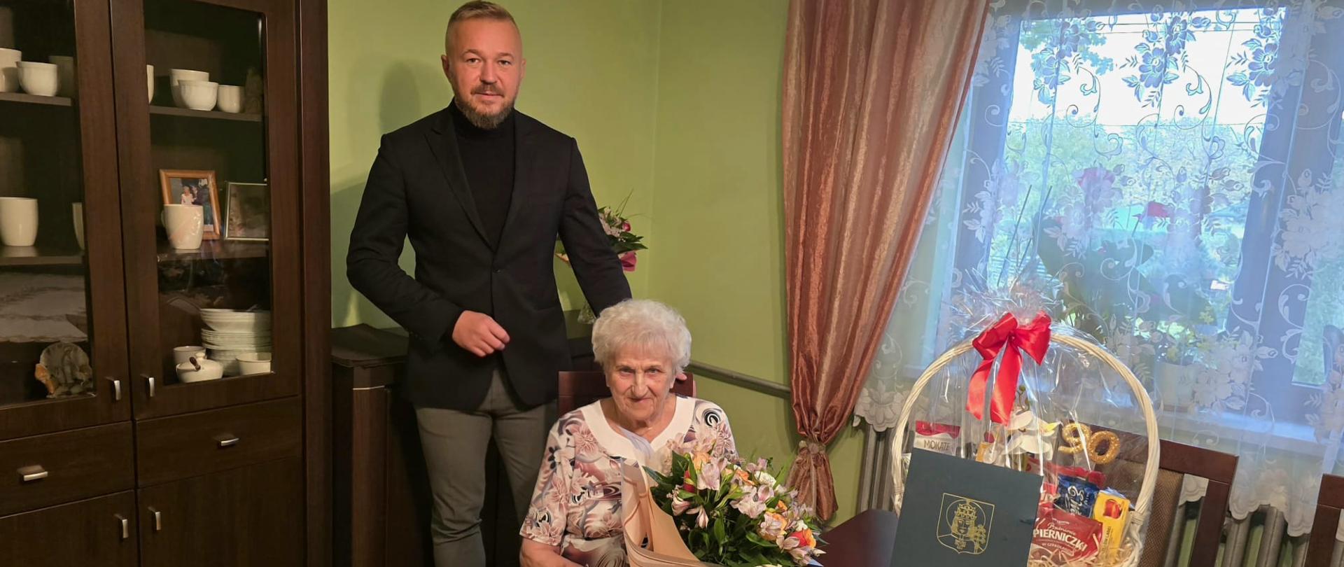 Wójt Gminy Kobiór odwiedził mieszkankę gminy z okazji jej jubileuszu. Seniorka siedzi przy stole z bukietem kwiatów, obok stoi wójt, a na stole znajduje się kosz upominkowy i pamiątkowa kartka z herbem Gminy Kobiór.