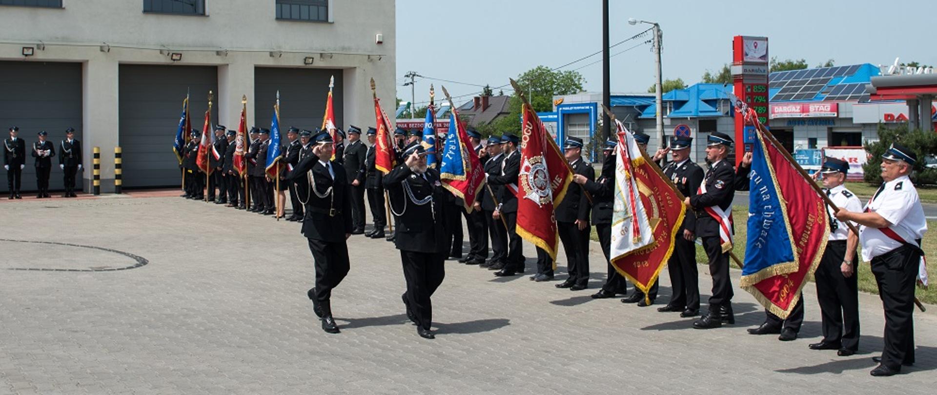 Dowódcy uroczystości przechodzą przez plac i salutują do ustawionych w szeregu pocztów sztandarowych jednostek OSP z terenu powiatu sokołowskiego. W tle ściana piętrowego budynku z czterema garażami i oknami. Na elewacji duży czerwony napis OSP "Cukrownia", herb miasta Sokołów Podlaski oraz rok założenia jednostki 1904