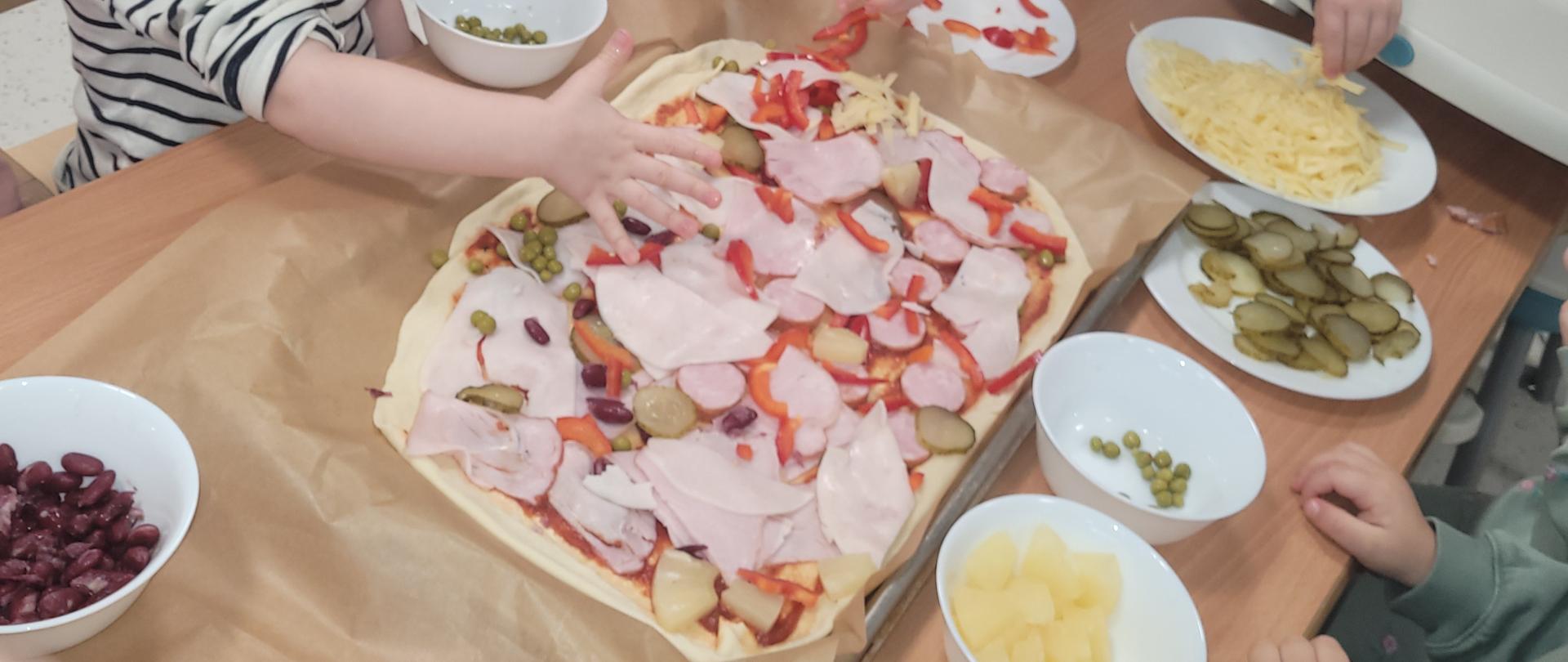 Zdjęcie w centralnej części przedstawia surowy spód pizzy, na którym dzieci w wieku około 2-2,5 lat układają poszczególne składniki takie jak wędlinę, ser, fasolę, ogórki kiszone i paprykę. Widać rączki dzieci. Poszczególne składniki są włożone w białe miseczki. Na twarzach dzieci widać skupienie. 