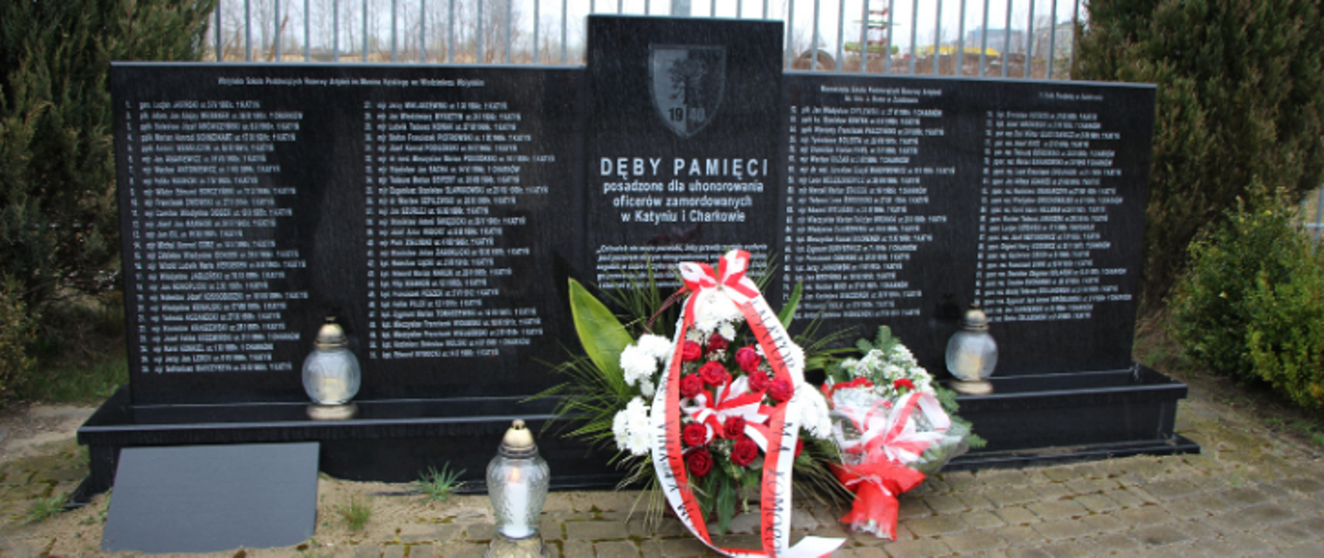 na zdjęciu znajduje się pomnik upamiętniający ofiary katyńskie przy ul. Ofiar Katynia w Zambrowie, a przy nim znajdują się biało-czerwone wiązanki kwiatów i 3 znicze, w tle zielone krzewy
