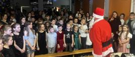 Na korytarzu szkolnym stoi Mikołaj. Przed nim stoją zgromadzeni: dzieci, rodzice i nauczyciele. Twarze wszystkich obecnych zwrócone są w stronę gościa.