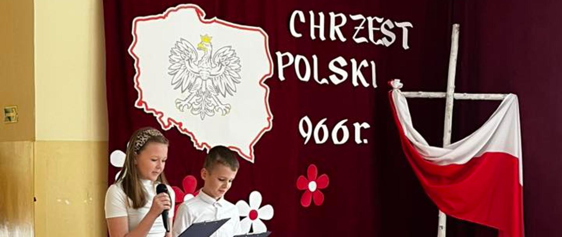 Dziewczynka i chłopiec prowadzący akademię przemawiają do mikrofonu, za nimi na bordowej zasłonie napis "Chrzest Polski 966 r." oraz wycięta z papieru mapa Polski z orłem w koronie pośrodku