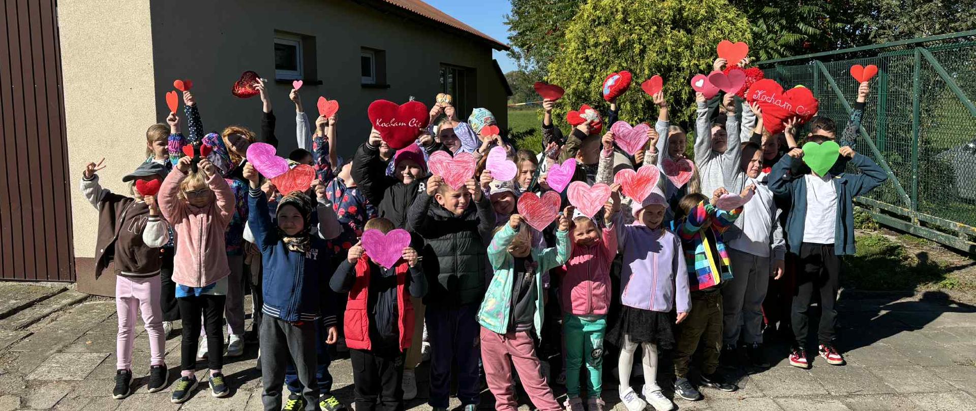 Na zdjęciu znajdują się uczniowie klas młodszych. Dzieci pozują do wspólnego zdjęcia. Wszyscy trzymają w dłoniach czerwone, papierowe serca.