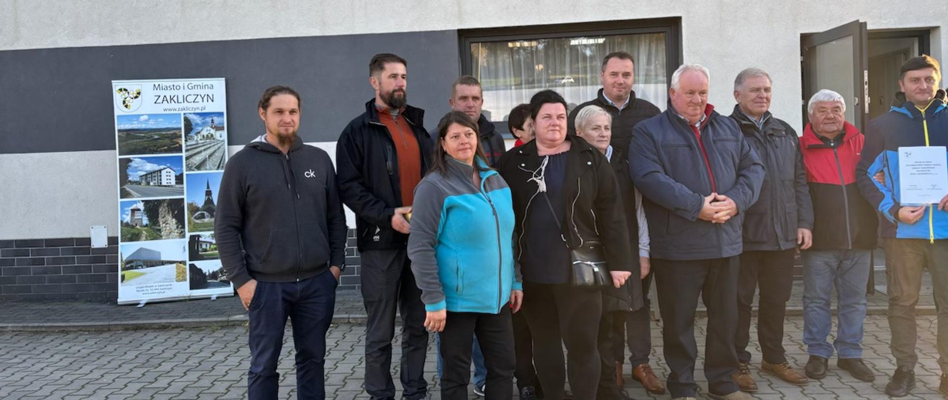 Grupa osób stoi przed budynkiem z napisem „Jedność Paleśnica”. W tle widoczny jest baner promujący Miasto i Gminę Zakliczyn. Część osób trzyma dokument, a cała grupa pozuje do wspólnego zdjęcia na zewnątrz.