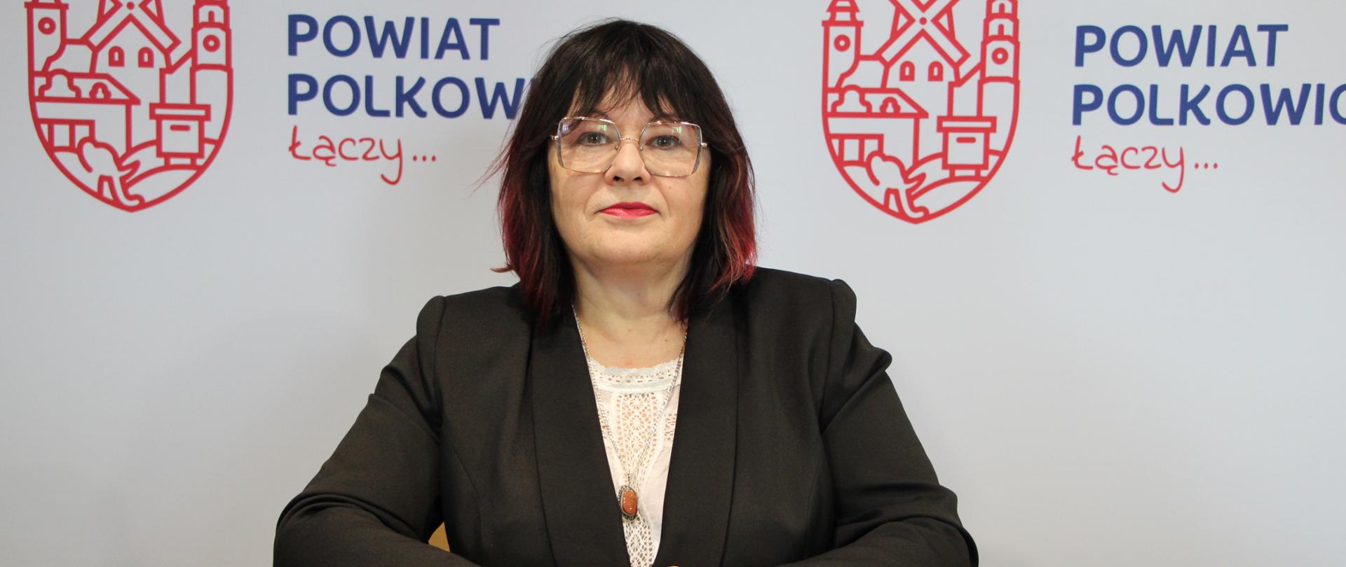 Kobieta w okularach siedzi przy stole , przed nią leży notes