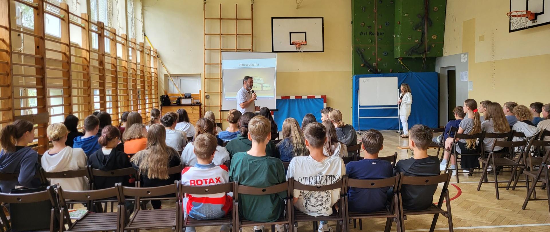 Dwoje dorosłych osób stoi przed uczniami siedzącymi na krzesłach na sali gimnastycznej.
