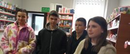 Dwie uczennice i dwóch uczniów stoi wewnątrz urzędu pocztowego, za nimi widoczne półki z towarami sprzedawanymi na poczcie