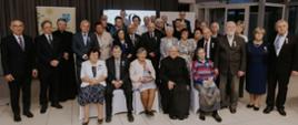 Duże, zbiorowe zdjęcie przedstawiające około 30 osób stojących i siedzących na tle ciemnej zasłony i ekranu z napisem "50 lat". Grupa składa się z odznaczonych par małżeńskich oraz urzędników i duchownych. Na pierwszym planie widać cztery osoby siedzące na krzesłach: dwie kobiety (jedna w białym żakiecie i czarnej spódnicy, druga w jasnoróżowym żakiecie) oraz dwóch mężczyzn (jeden w garniturze, drugi w czarnej sutannie). Większość osób w drugim i trzecim rzędzie stoi, a większość odznaczonych mężczyzn ma przypięte medale do garniturów. Ubrane są głównie w eleganckie, ciemne lub jasne stroje. Po lewej stronie widoczny jest baner Gminy Siepraw.
