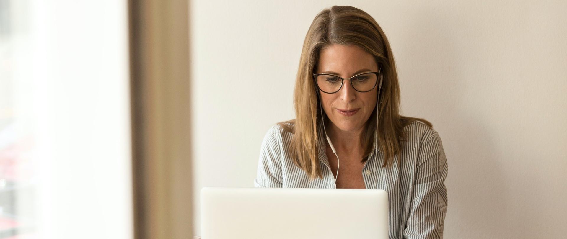 Kobieta w okularach i słuchawkach na uszach siedzi przy biurku i pisze na laptopie. Obok leży telefon i gazeta.
