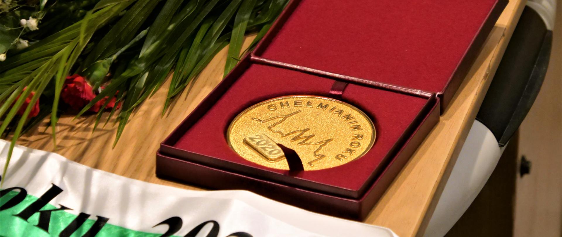 Zdjęcie przedstawia złoty medal leżący na stole w etui. Przed medalem leży szarfa z napisem "Chełmianin Roku 2020"