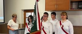 W centrum kadru znajduje się trzyosobowy poczet sztandarowy złożony z uczniów w strojach galowych (białe koszule, ciemne spodnie), z biało-czerwonymi szarfami i w białych rękawiczkach.