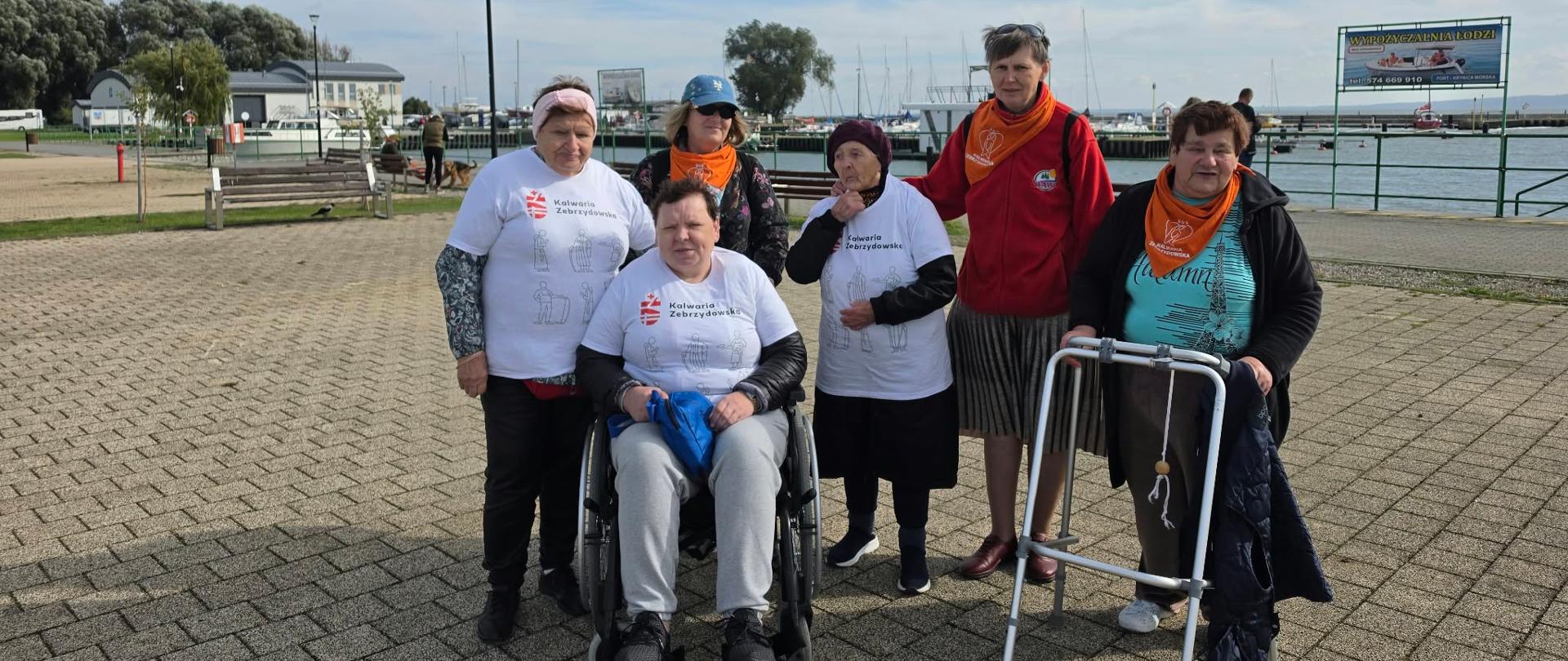 Zdjęcie grupowe sześciu kobiet, w tym osób z niepełnosprawnościami, podczas wyjazdu nad morze. Uczestniczki pozują na nadmorskiej promenadzie, w pobliżu portu i zacumowanych łodzi. Część kobiet ma na sobie koszulki z logo Kalwarii Zebrzydowskiej. Jedna z uczestniczek porusza się na wózku inwalidzkim, a inna korzysta z balkonika. W tle widoczne są łodzie, zabudowania portowe i morze.