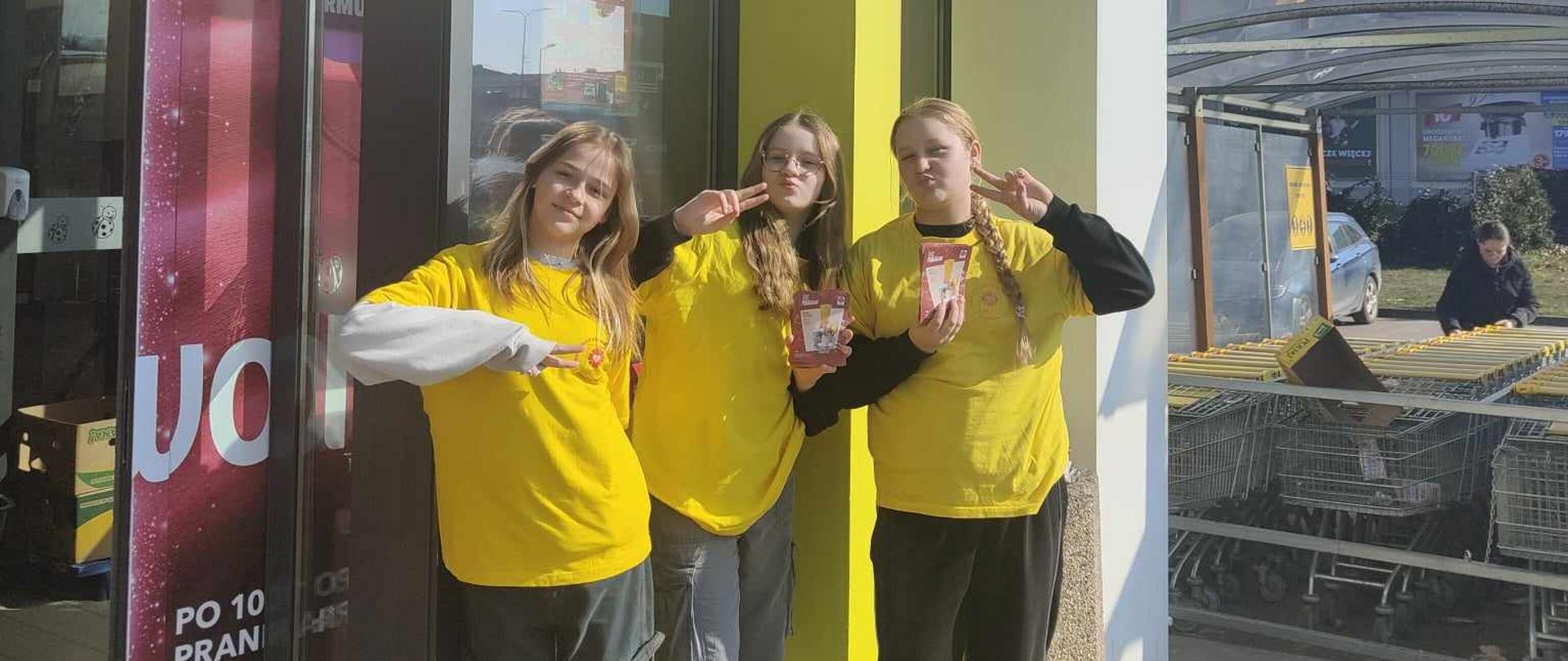 Trzy dziewczyny stojące pod drzwiami do marketu, mają żółte koszulki, trzymają ulotki, podnoszą palce do góry układając je w kształt v.