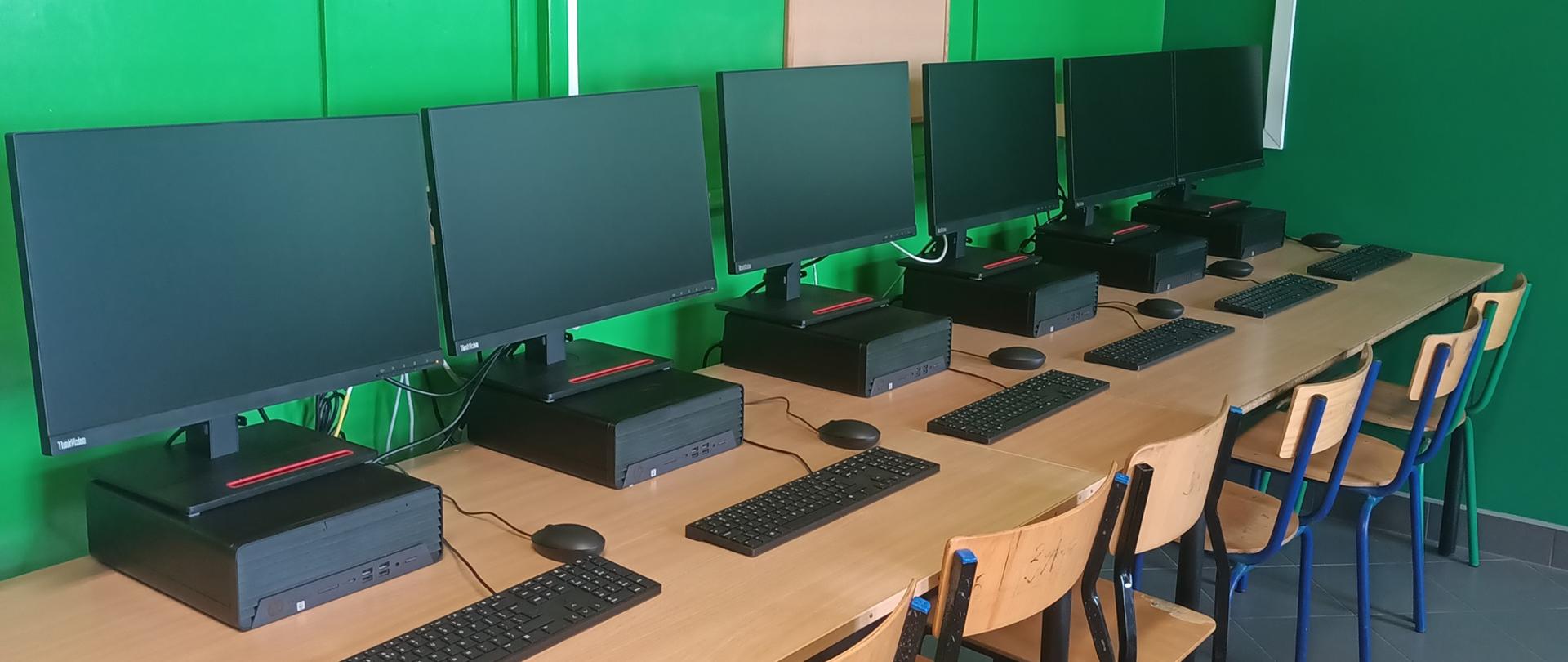 Połączone biurka przy ścianie. Na nich stoją komputery z monitorami a przy nich krzesła