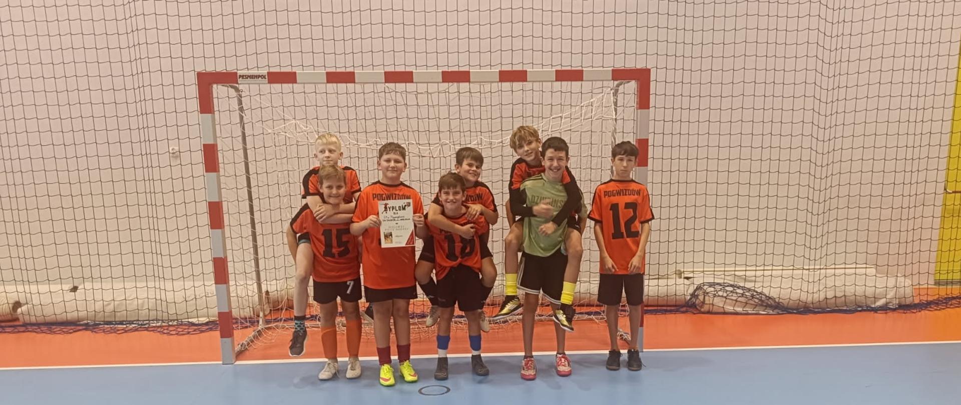 Ośmiu uśmiechniętych chłopców w jednakowych pomarańczowych podkoszulkach i czarnych spodenkach soi na hali sportowej na tle bramki piłkarskiej. Jeden z chłopaków w ręce trzyma dyplom.