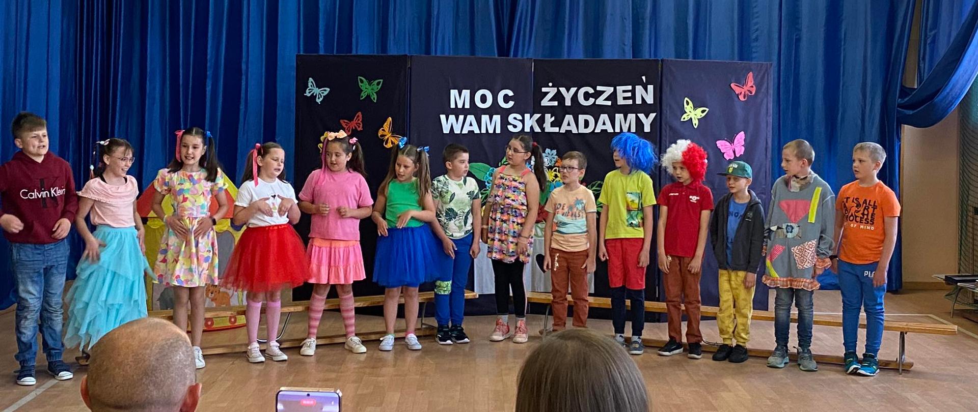 Dzieci poubierane w różnokolorowe ubrania stoją obok siebie tle niebieskiej kotary z napisem Moc życzeń wam składamy
