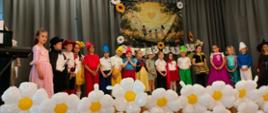 dzieci przebrane w stroje bajkowe stoją na scenie ozdobionej dmuchanymi kwiatami