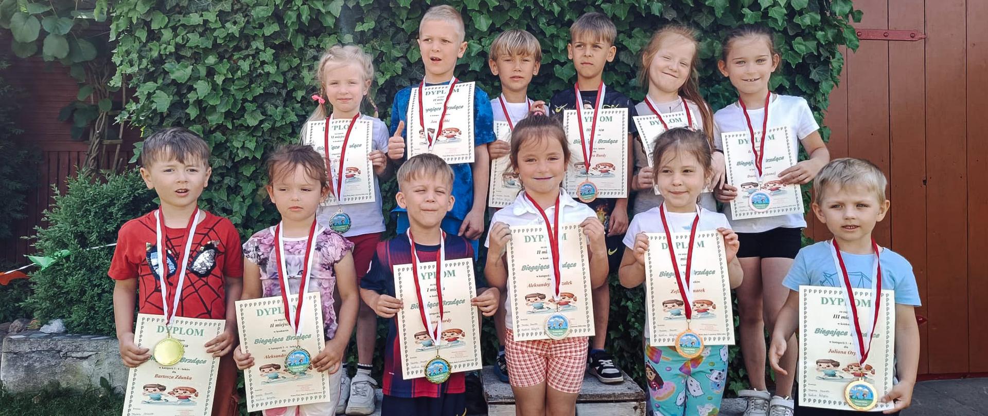 Grupa dzieci w wieku przedszkolnym ustawiona w dwóch rzędach. Drugi rząd dzieci stoi na podwyższeniu, na białych odwróconych skrzyniach. Wszyscy maja zawieszone na szyjach medale a w rękach trzymają dyplomy. Za nimi widać ścianę pokrytą zielenią oraz drzwi. 
