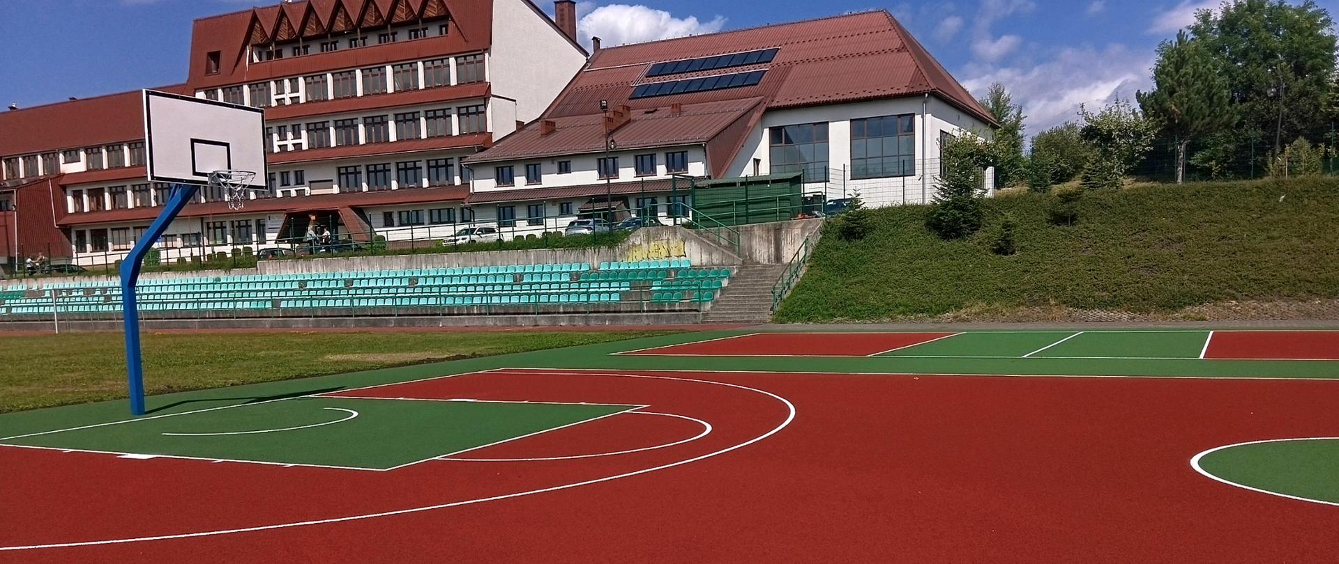 Kompleks sportowy w Skawie – wielofunkcyjne boisko o nawierzchni syntetycznej w kolorze czerwonym i zielonym, z wyznaczonymi liniami boiskowymi i koszem do koszykówki. W tle widoczne są trybuny z siedziskami oraz budynki szkolne.