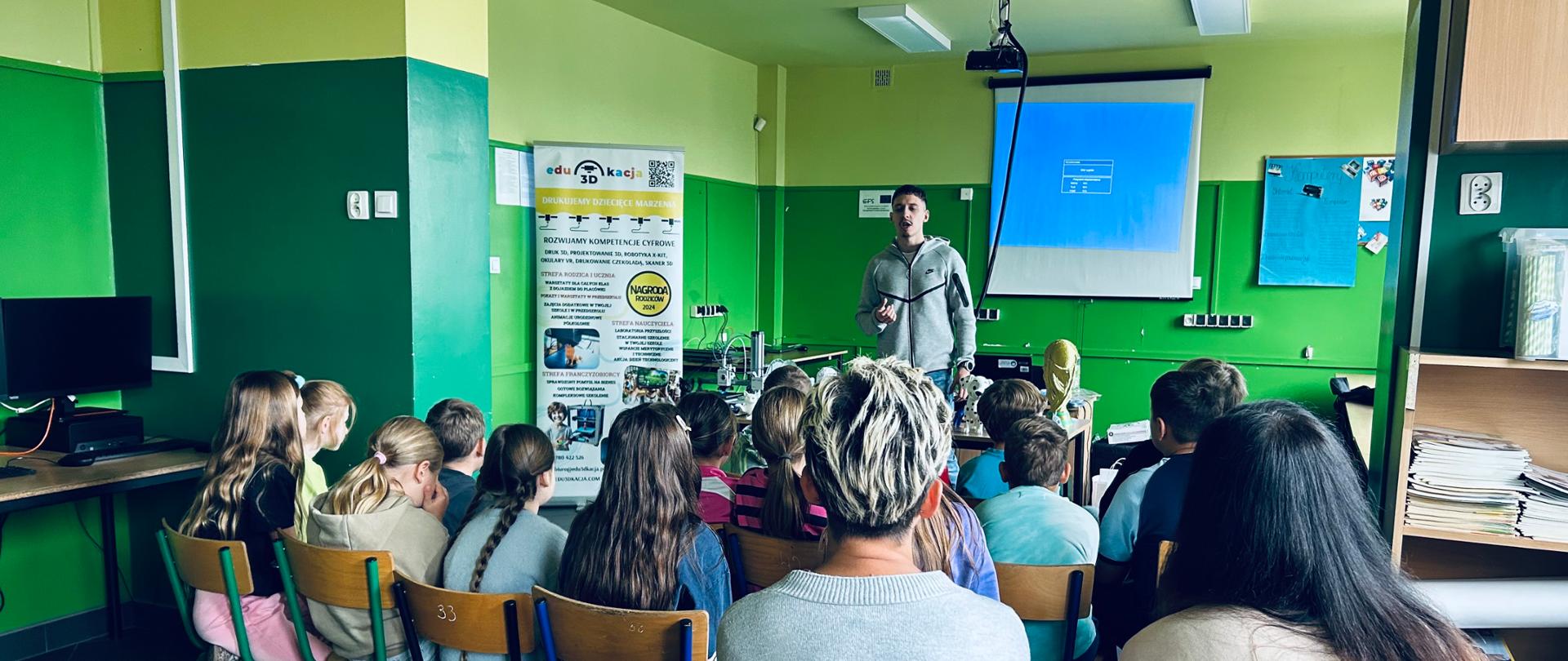 Uczniowie w szkolnej pracowni komputerowej siedzą tyłem do aparatu na drewnianych krzesłach i słuchają prowadzącego, który stoi przed ekranem projekcyjnym. Obok niego stoi roll-up edu3Dkacja, a na stole za nim widać wydruki 3D. Ściany sali są jasnozielone i żółte.