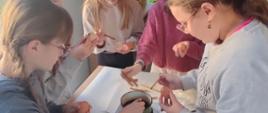 Dziewczynki z klasy 5 pochylają się nad stołem w bibliotece, każda trzyma w ręku jajko, w drugiej ręce pędzelek, który zanurzają w garnku z woskiem