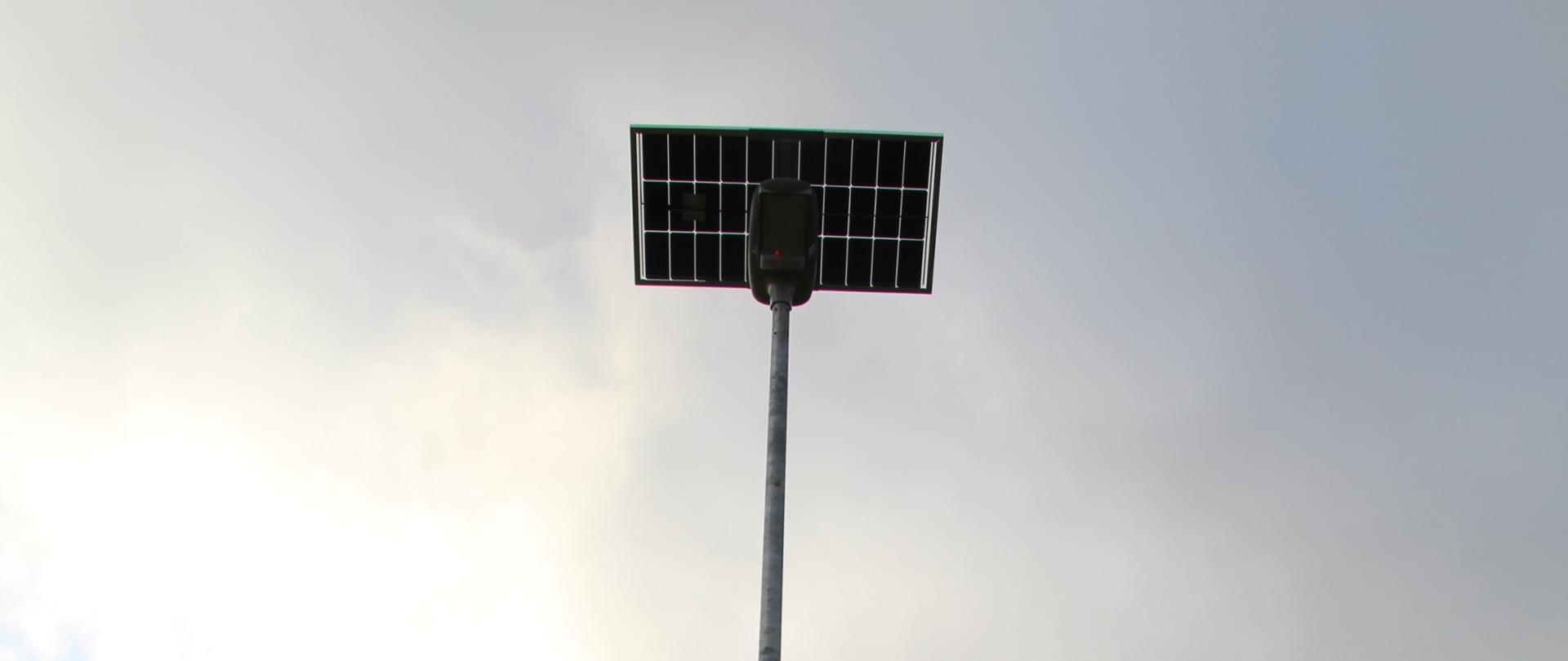 Latarnia solarna o srebrnym słupie i panelu fotowoltaicznym na szczycie, umieszczona na środku zdjęcia. Latarnia stoi na porośniętym trawą terenie, prawdopodobnie na skraju pola lub łąki. Po prawej stronie widoczne są wysokie, ciemnozielone krzewy iglaste. W tle, na horyzoncie, rozciąga się otwarty teren z pojedynczymi drzewami. Niebo jest zachmurzone, z wyraźnym prześwitem słońca po lewej stronie. Na słupie widoczna jest niewielka, biała tablica informacyjna.