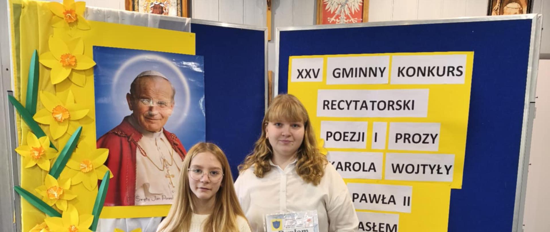 Dwie uczennice ubrane w stroje galowe pozują z dyplomami. Za nimi na tablicy widnieje zdjęcie Karola Wojtyły oraz napis: Dwudziesty piąty Gminny Konkurs Recytatorski Poezji i Prozy Karola Wojtyły "Naprzód z Nadzieją".